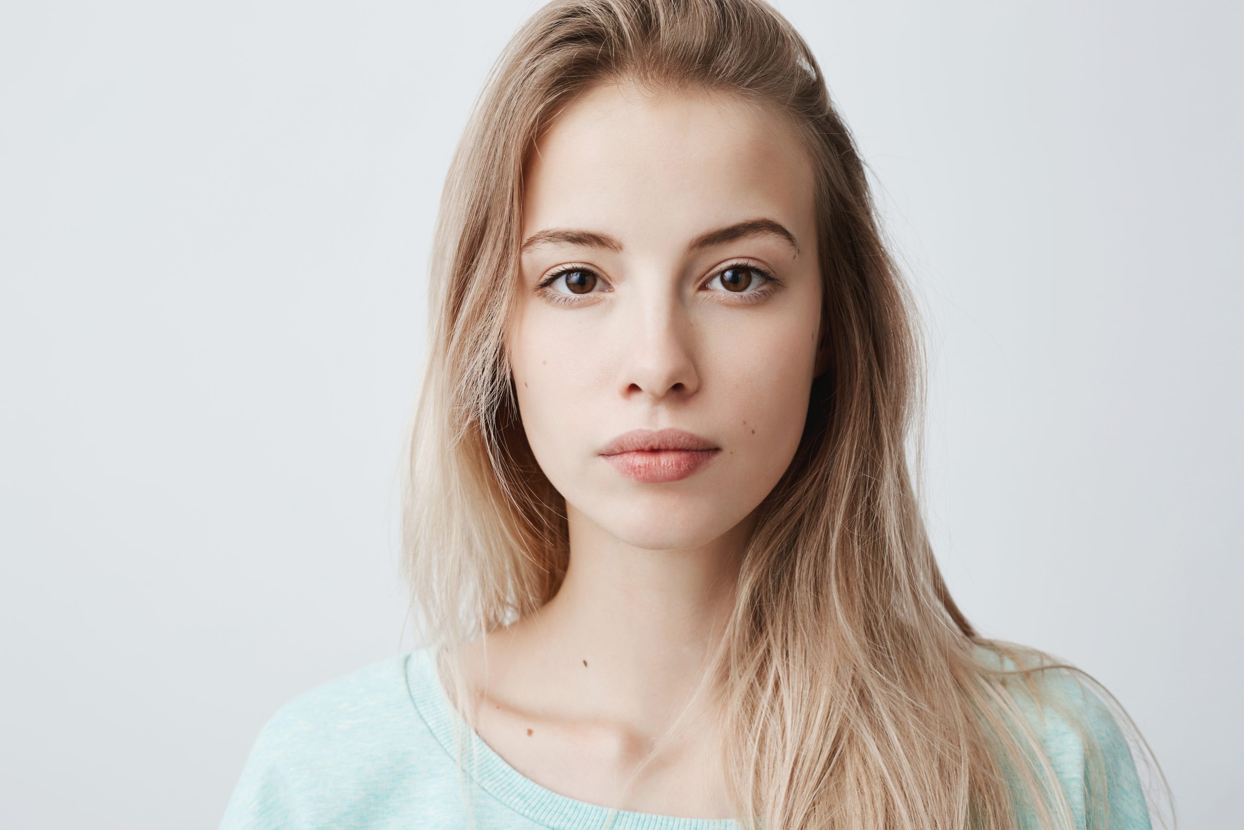 Portrait of fair-haired woman with warm dark eyes, healthy skin looking directly at camera with pleased and calm expression. Alluring girl with beautiful appearance dressed casually posing at studio. Beauty concept